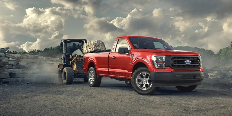 Pictured is a brand new red 2023 Ford F-150 towing large construction equipment through a construction site.