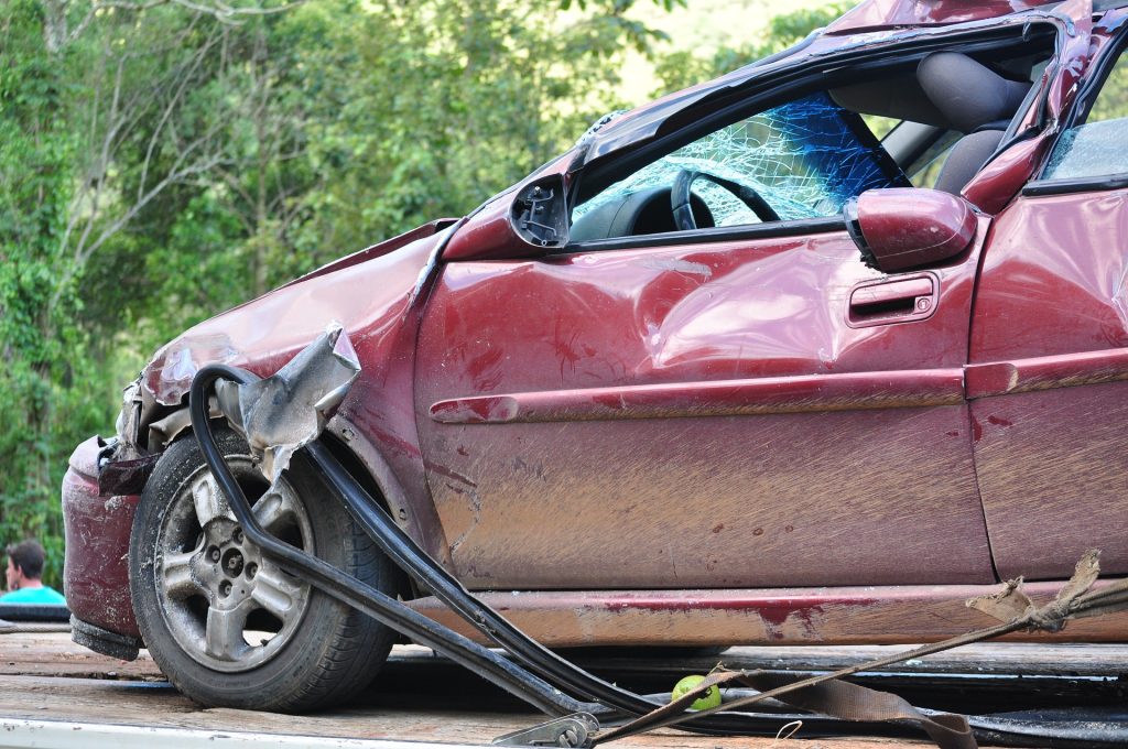 red vehicle on the back of a tow truck that looks completely totaled. Road way side rails are sticking out of the bottom of the vehicle and the front is smashed in.