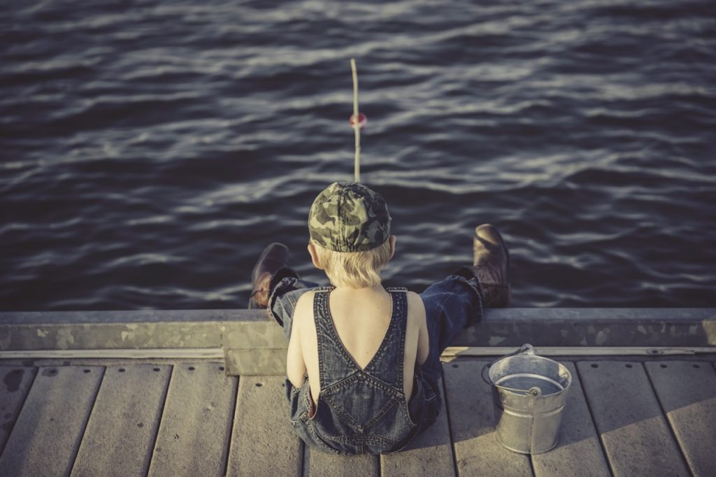 little boy sitting on the edge of a dock in his overalls with no shirt and a camo ball cap fishing.